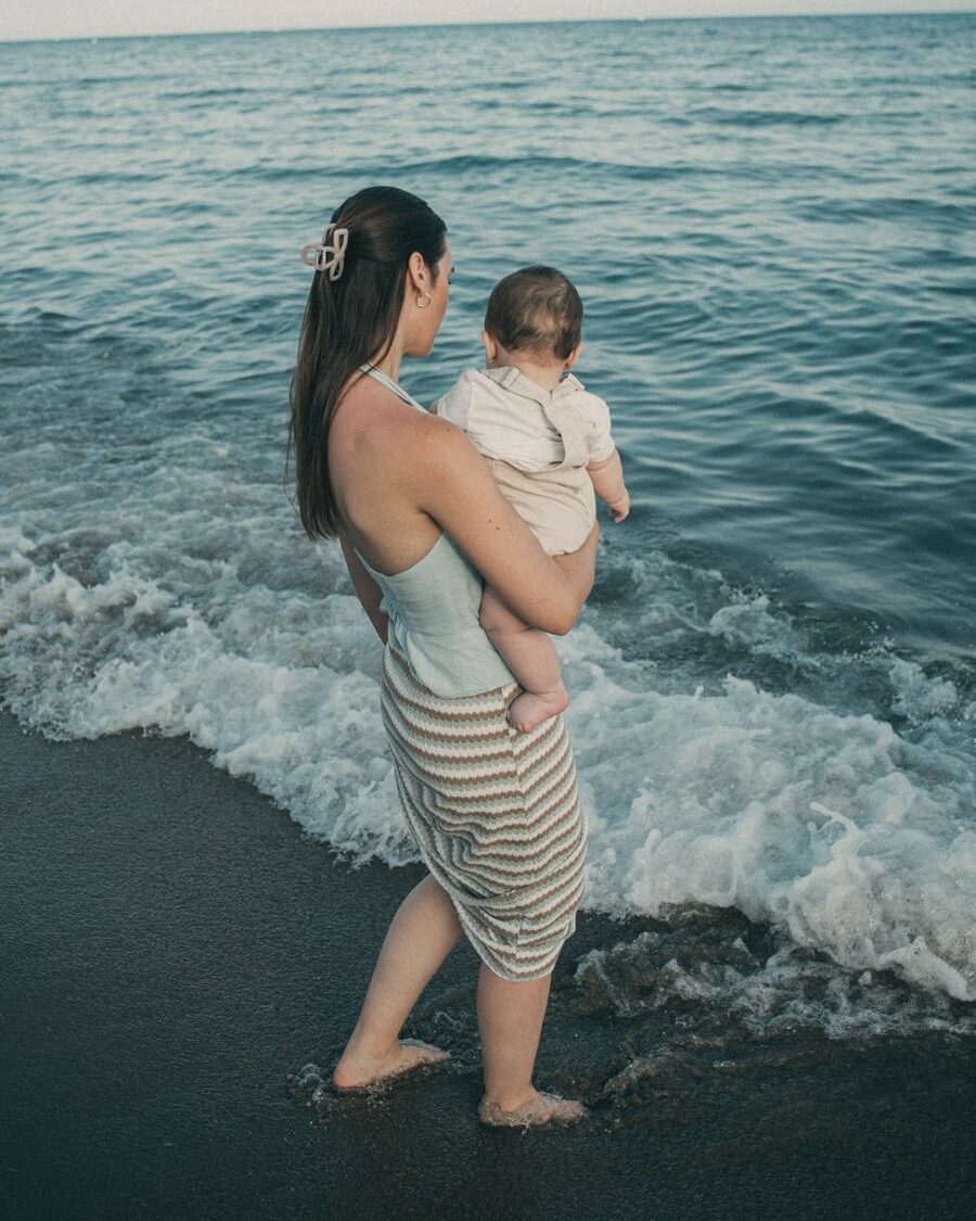 Myriam en la orilla de la playa con su bebé en brazos, mirando las olas