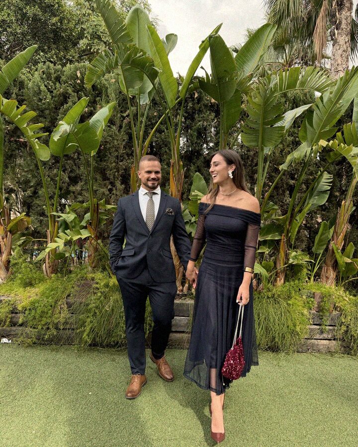 Myriam y su pareja de la mano, riendo, en un jardín con plataneras